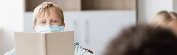 Colegial en máscara médica mirando a la cámara mientras sostiene el libro en el aula, pancarta - foto de stock