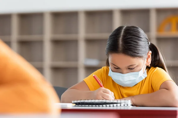 Asiático colegiala en médico máscara escritura en notebook en aula - foto de stock