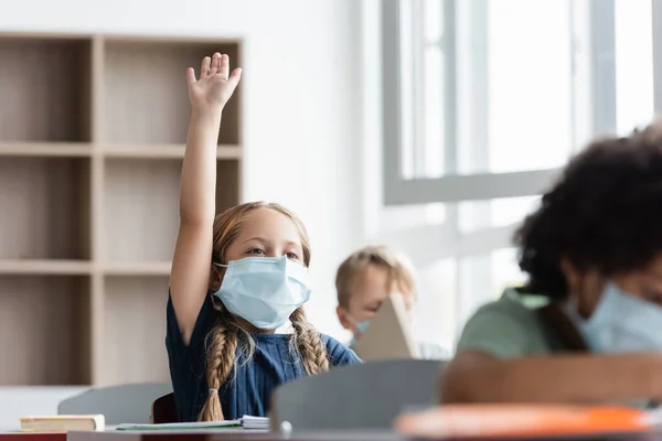 Écolière en masque médical levant la main pendant la leçon près de camarades de classe multiculturels — Photo de stock