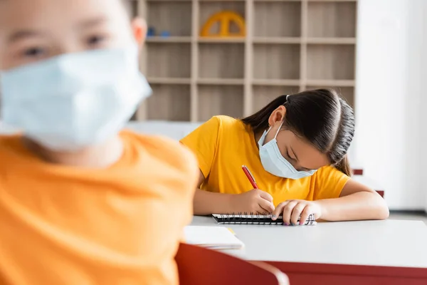 Asiatique écolière dans médical masque écriture dans notebook pendant leçon — Photo de stock