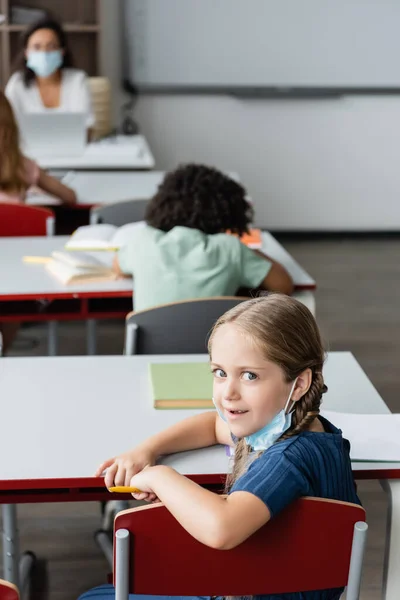 Écolière avec masque médical sur le menton regardant la caméra dans la salle de classe — Photo de stock