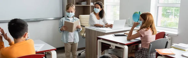 Boy in medical mask reading book near african american teacher and pupils in classroom, banner — Stock Photo