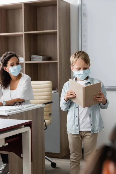 Schoolboy in medical mask reading book near african american teacher during lesson — Stock Photo