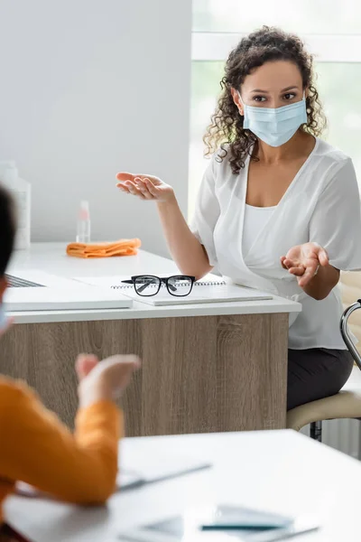 African american teacher in medical mask pointing with hands near blurred pupil — Stock Photo