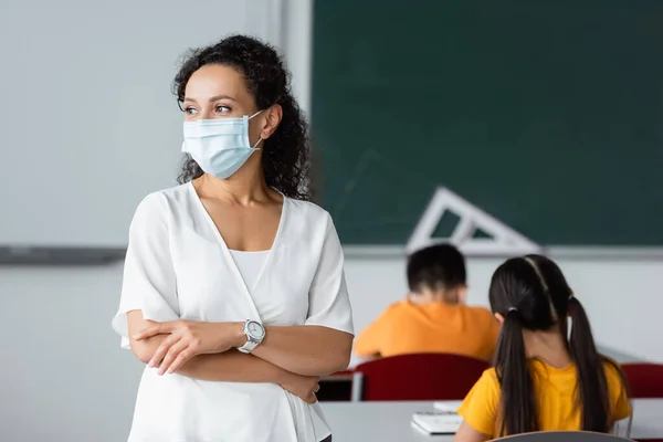 Profesor afroamericano en máscara médica de pie con los brazos cruzados en el aula - foto de stock