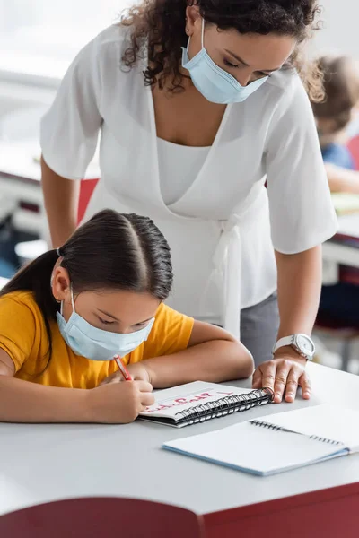 Africano americano maestro en médico máscara de pie cerca asiático chica escritura en notebook - foto de stock