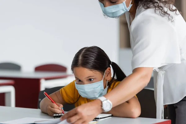 Afro-américain professeur dans masque médical près asiatique fille écriture dans notebook — Photo de stock
