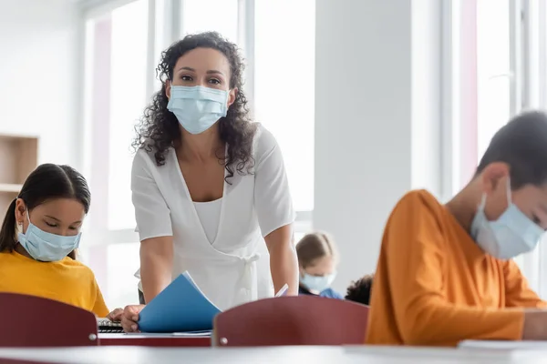 African american teacher in medical mask near multiethnic kids in classroom — Stock Photo