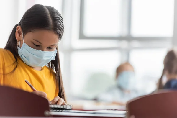 Asian schoolgirl in medical mask writing in notebook in classroom — Stock Photo