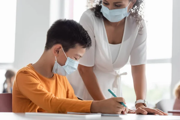 Африканский учитель-американец в медицинской маске стоит рядом с азиатским мальчиком в классе — стоковое фото