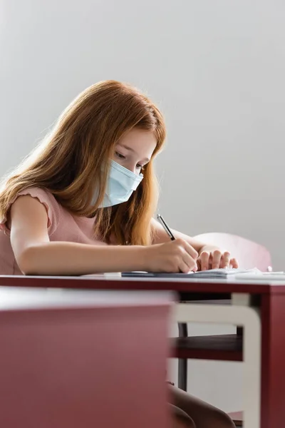 Écolière en masque médical écrivant pendant les cours à l'école — Photo de stock