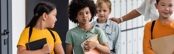 Pensive african american schoolboy holding notebooks near multiethnic pupils and teacher, banner — Stock Photo