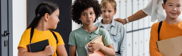 Pensativo afroamericano colegial sosteniendo cuadernos cerca de alumnos multiétnicos y maestro, pancarta - foto de stock