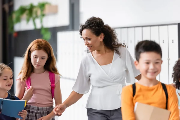 Sorridente insegnante afro-americana che cammina nel corridoio scolastico con bambini multietnici — Foto stock