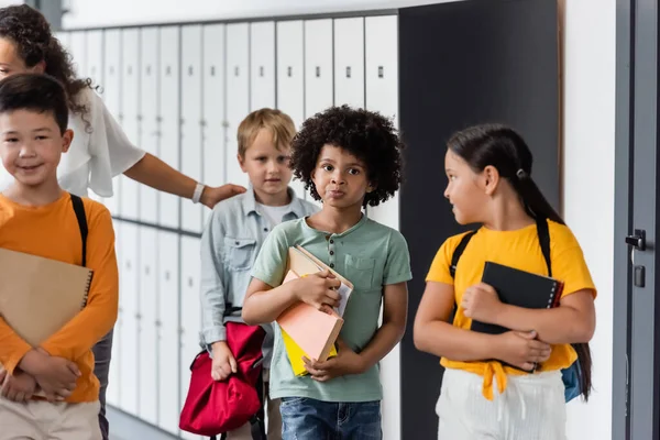 Reflexivo afroamericano chico hinchando las mejillas cerca de compañeros de clase multiétnicos y maestro en la sala de la escuela - foto de stock