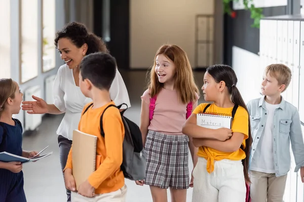 Fröhliche afrikanisch-amerikanische Lehrerin zeigt mit der Hand, während sie mit multiethnischen Kindern auf dem Schulflur spricht — Stockfoto