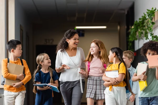 Profesor afroamericano positivo gesticulando mientras habla con alumnos multiculturales en el salón de la escuela - foto de stock