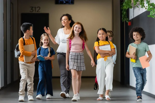 Fröhliche multiethnische Schüler gehen mit afrikanisch-amerikanischem Lehrer den Schulflur entlang — Stockfoto