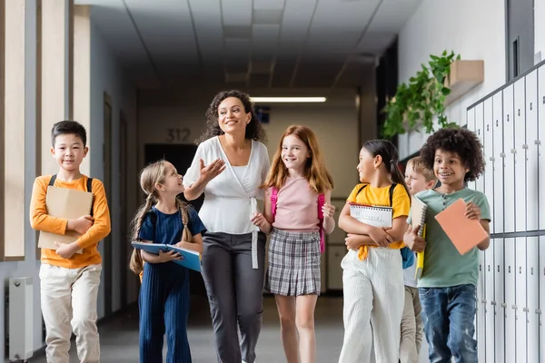 Professeur afro-américain excité parlant à des enfants multiethniques heureux dans le hall de l'école — Photo de stock