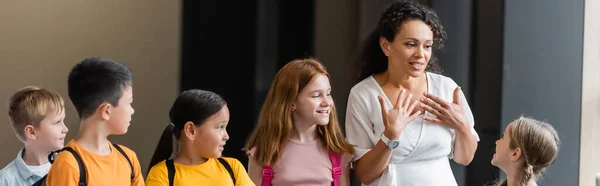 Enseignant afro-américain parlant à des enfants multiculturels positifs dans la salle d'école, bannière — Photo de stock