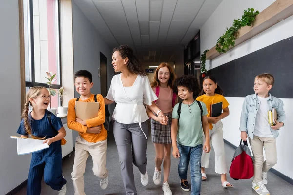 Schüler mit Notizbüchern laufen mit afrikanisch-amerikanischem Lehrer in der Schulaula — Stockfoto