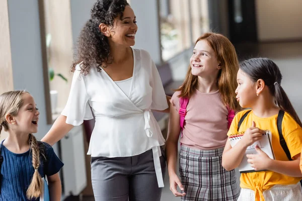 Fröhliche multiethnische Schulmädchen zu Fuß in Korridor nahe afrikanisch-amerikanischer Lehrerin — Stockfoto