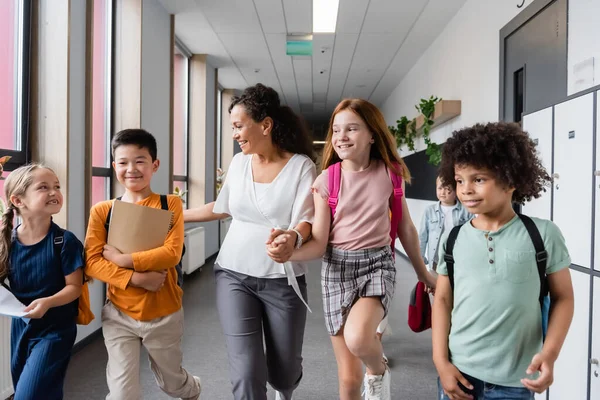 Allegri alunni multiculturali che camminano lungo il corridoio scolastico con insegnante afro-americano — Foto stock