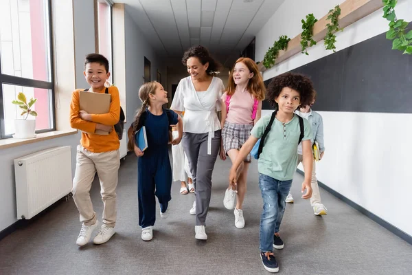 Happy multiethnic kids and african american teacher walking in school corridor — Stock Photo