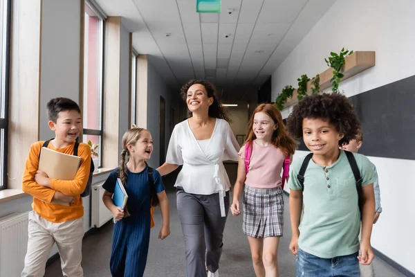 Allegri bambini multiculturali che camminano nel corridoio scolastico con insegnante afro-americano — Foto stock