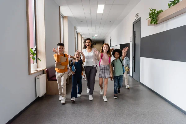 Felici alunni multiculturali con insegnante afro-americano che cammina nel corridoio scolastico e agita le mani — Foto stock