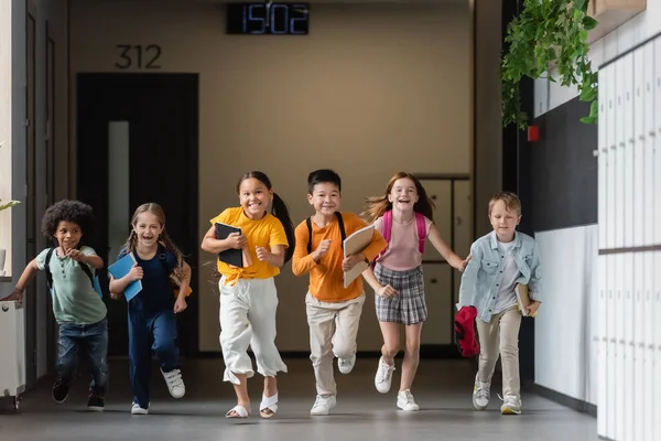 Alegres amigos multiculturales con cuadernos corriendo en el pasillo de la escuela - foto de stock