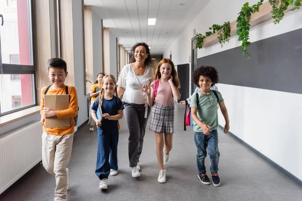 Glücklich afrikanisch-amerikanischer Lehrer läuft mit multiethnischen Schülern auf dem Schulflur — Stockfoto