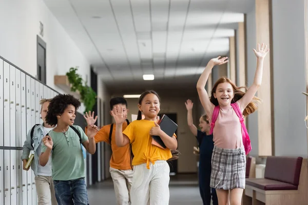 Happy multiethnic pupils waving hands while running in school corridor — Stock Photo