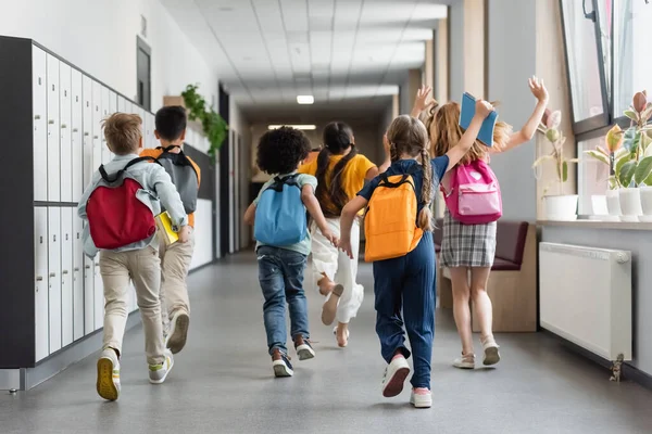 Vista trasera de los niños interracial con mochilas agitando las manos mientras se ejecuta en el pasillo de la escuela - foto de stock