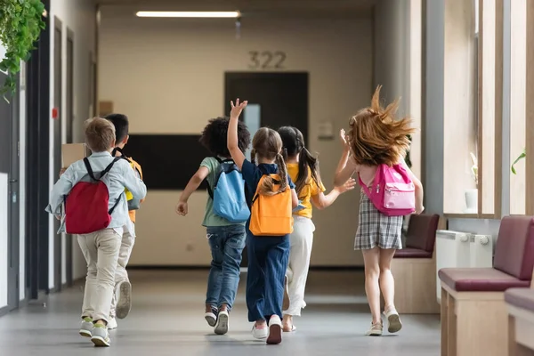 Vue arrière d'amis multiethniques avec des sacs à dos courant et agitant les mains dans le couloir de l'école — Photo de stock