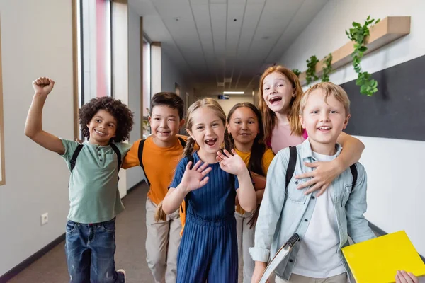 Fröhliche multiethnische Kinder, die sich in der Schule umarmen, Hände winken und Jubelgeste zeigen — Stockfoto