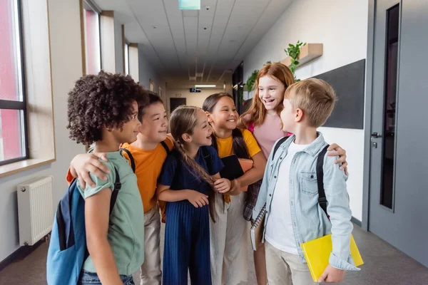 Crianças multiculturais alegres conversando e abraçando no salão da escola — Fotografia de Stock