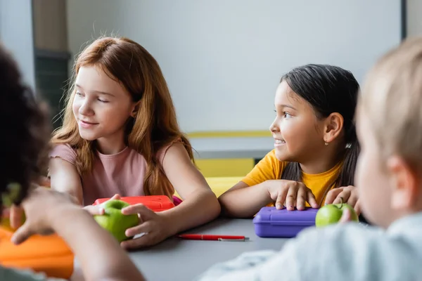 Interracial schoolgirls near lunch boxes and blurred friends in school eatery — Stock Photo