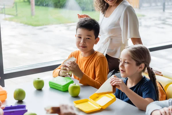 Felice asiatico ragazzo holding sandwich durante pranzo vicino studentessa e africano americano insegnante — Foto stock