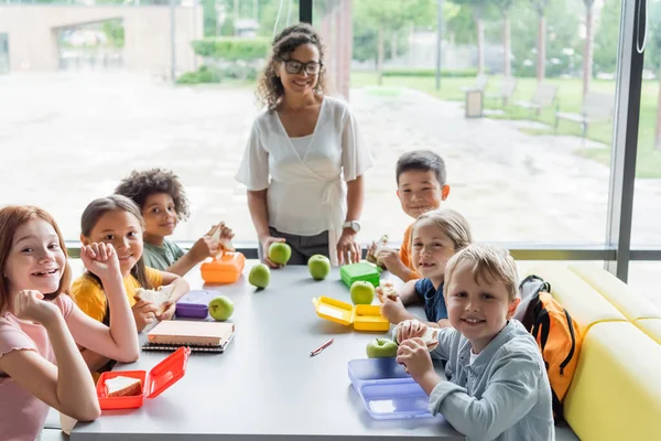 Felizes estudantes multiétnicos olhando para a câmera perto de professor afro-americano no restaurante da escola — Fotografia de Stock