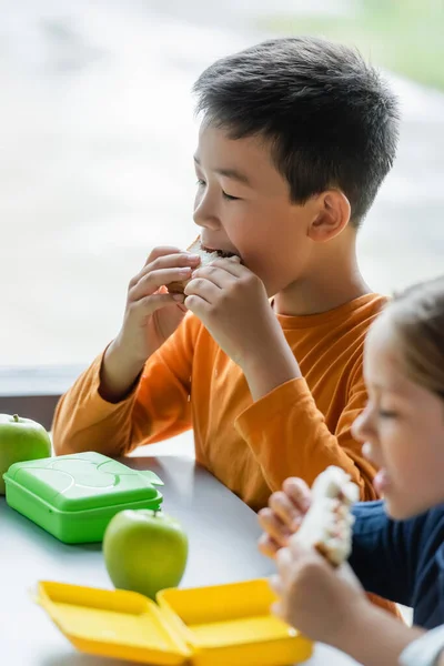 Азіатський школяр їсть бутерброд біля розмитої дівчини в шкільному харчуванні — стокове фото