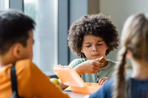 African american boy eating sandwiches near blurred classmates — Stock Photo