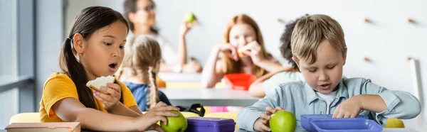 Asian schoolgirl eating sandwich near classmates during lunch break, banner — Stock Photo