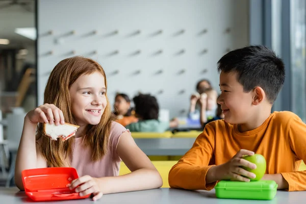 Redhead girl with sandwich smiling to asian boy in school dining room — Stock Photo