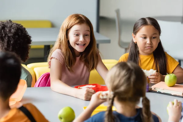 Збуджена руда дівчина посміхається біля міжрасових однокласників, обідаючи в їжі — стокове фото