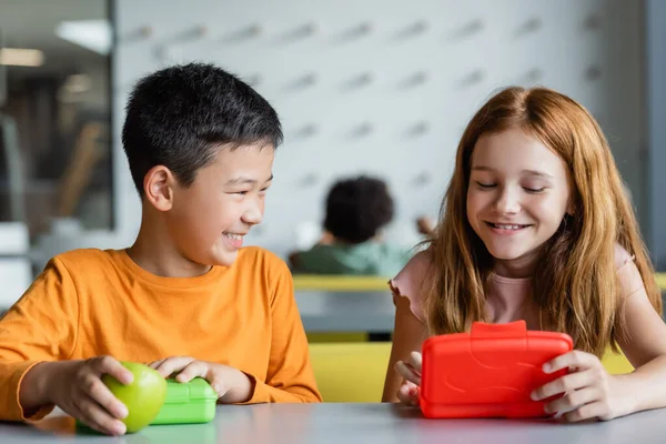 Rousse fille et sourire asiatique garçon près de déjeuner boîtes dans école salle à manger — Photo de stock