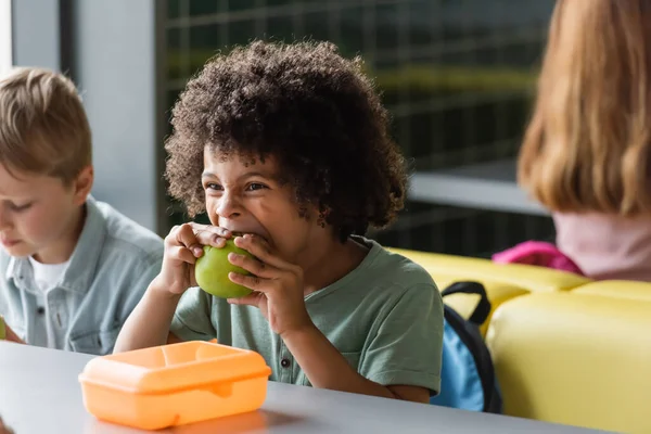 Африканский американский школьник ест яблоко рядом с размытыми одноклассниками в школьной столовой — стоковое фото