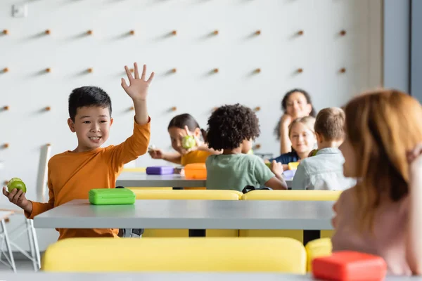 Cheerful asian boy waving hand to blurred girl in school eatery — Stock Photo