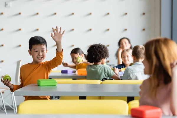 Alegre asiático chico agitando mano a borrosa chica en escuela eatery - foto de stock