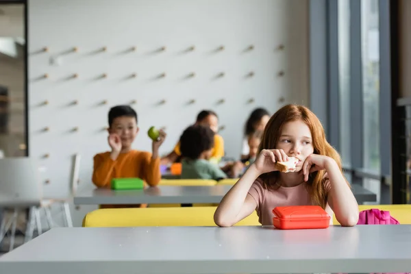 Triste rousse fille manger sandwich près des écoliers sur fond flou — Photo de stock